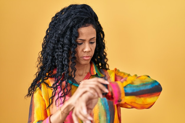 Middle age hispanic woman standing over yellow background checking the time on wrist watch relaxed and confident