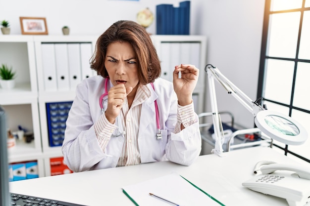 Middle age hispanic woman wearing doctor uniform and stethoscope at the clinic feeling unwell and coughing as symptom for cold or bronchitis. health care concept.