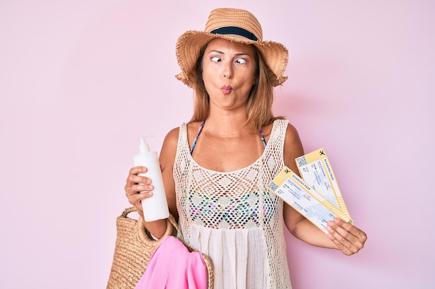 Middle age hispanic woman wearing summer hat holding boarding pass and sunscreen making fish face with mouth and squinting eyes crazy and comical