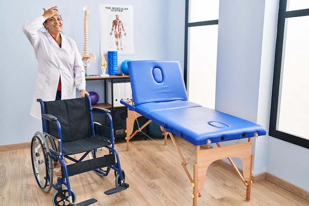 Middle age hispanic woman with wheelchair at physiotherapy clinic stressed and frustrated with hand on head, surprised and angry face