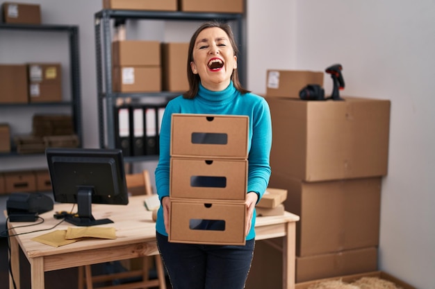 Middle age hispanic woman working at small business ecommerce holding boxes smiling and laughing hard out loud because funny crazy joke