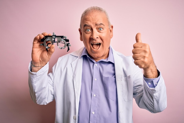 Middle age hoary optical man wearing coat holding optometry glasses over pink background screaming proud and celebrating victory and success very excited cheering emotion