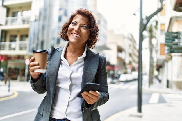 Middle age latin businesswoman drinking coffee and using smartphone at the city.