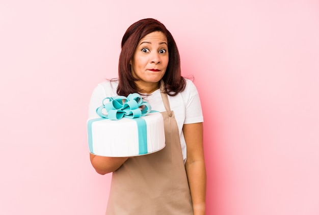 Middle age latin woman holding a cake isolated on a pink wall shrugs shoulders and open eyes confused.