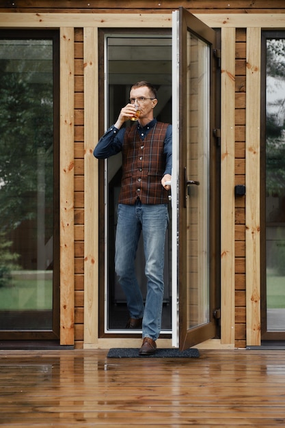 Middle age man comes out on the terrace of a small wooden house with a glass in hand