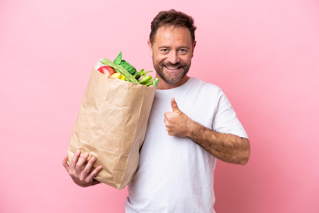 Middle age man holding a grocery shopping bag isolated on pink background giving a thumbs up gesture