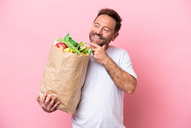 Middle age man holding a grocery shopping bag isolated on pink background looking up while smiling