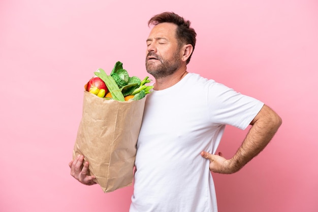 Middle age man holding a grocery shopping bag isolated on pink background suffering from backache for having made an effort