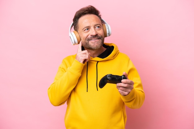 Middle age man playing with a video game controller isolated on pink background thinking an idea while looking up