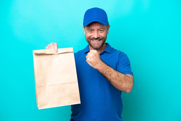 Middle age man taking a bag of takeaway food isolated on blue background celebrating a victory