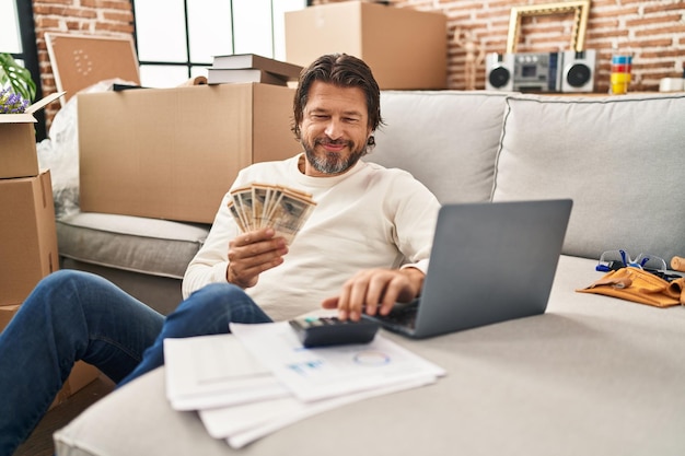 Middle age man using laptop counting denmark kroner banknotes at new home