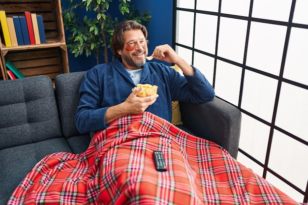 Middle age man wearing under eye patches watching movie eating chips potatoes at home