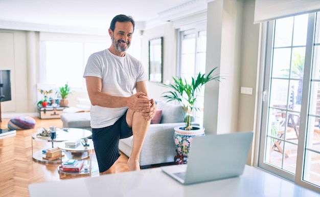 Middle age man with beard training and stretching doing exercise at home looking at sport video on computer