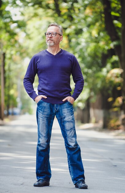 Middle age man with gray hair in park. Blue sweater. Eyeglasses. Bearded confident businessman. Blurred background. Hands in pockets. Half size photo