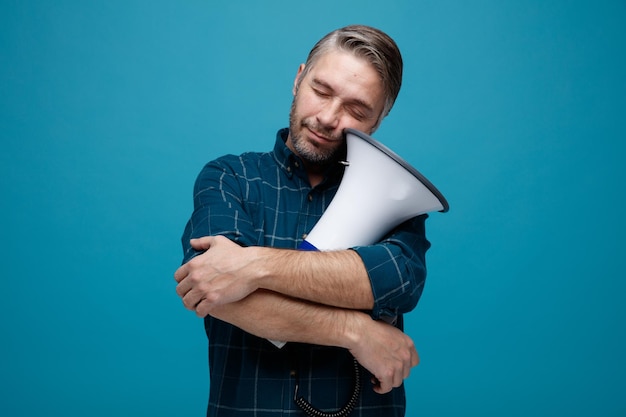 Middle age man with grey hair in dark color shirt holding megaphone looking happy and pleased standing over blue background