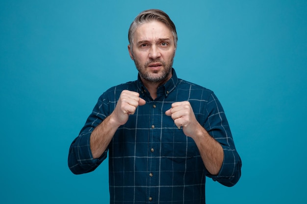 Middle age man with grey hair in dark color shirt looking at camera with serious tense face clenching fists ready to fight standing over blue background