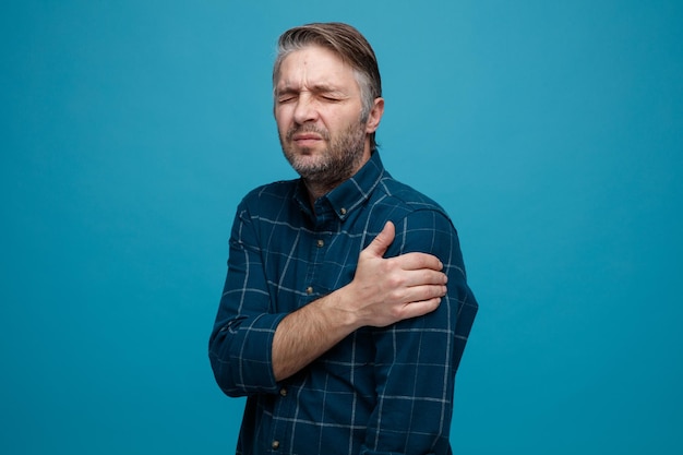 Middle age man with grey hair in dark color shirt looking unwell touching his shoulder feeling pain standing over blue background