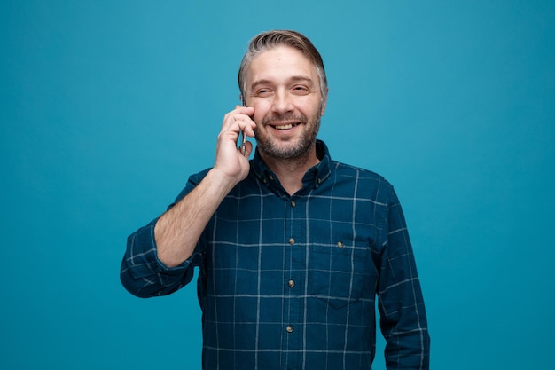 Middle age man with grey hair in dark color shirt talking on mobile phone smiling cheerfully happy and positive standing over blue background