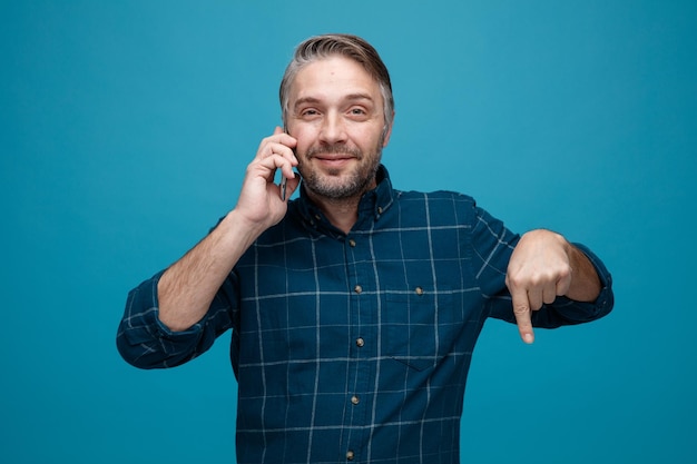 Middle age man with grey hair in dark color shirt talking on mobile phone smiling pointing with index finger down standing over blue background