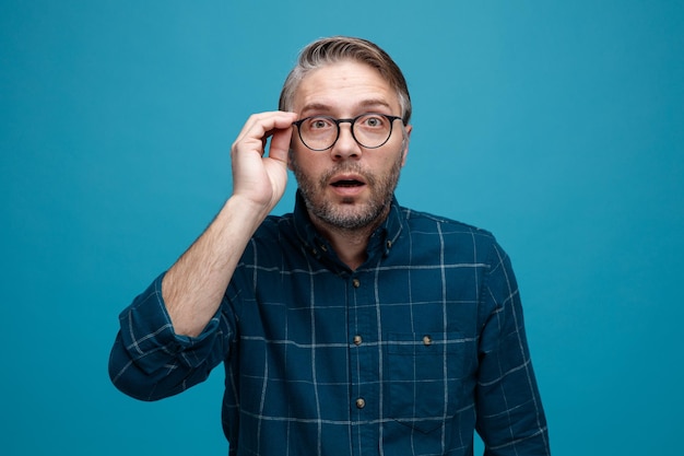 Middle age man with grey hair in dark color shirt wearing glasses staring at camera being surprised standing over blue background