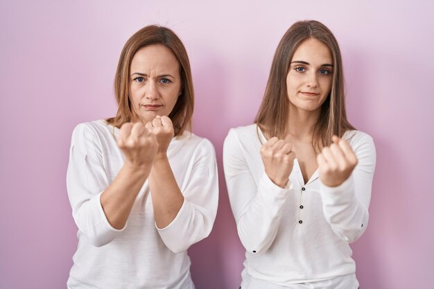 Middle age mother and young daughter standing over pink background ready to fight with fist defense gesture angry and upset face afraid of problem