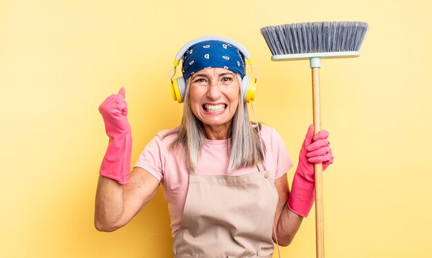 Middle age pretty woman shouting aggressively with an angry expression. household and broom concept