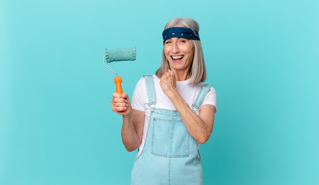 Middle age white hair woman feeling happy and facing a challenge or celebrating with a roller painting a wall