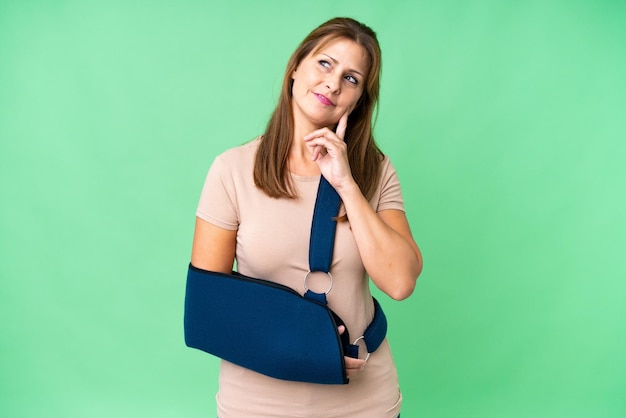 Middle age with broken arm and wearing a sling over isolated background thinking an idea while looking up