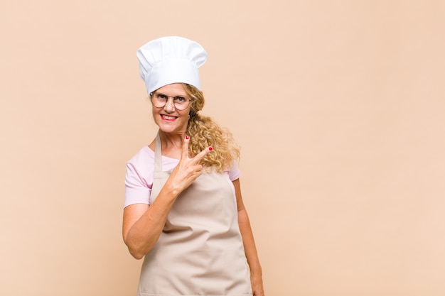 Middle age woman baker feeling happy, positive and successful, with hand making v shape over chest, showing victory or peace