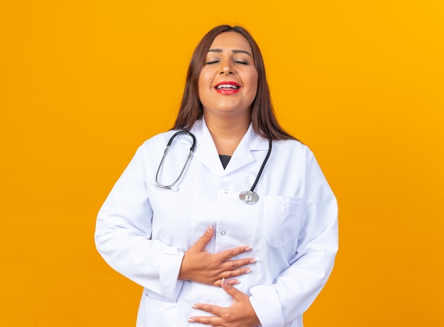 Middle age woman doctor in white coat with stethoscope happy and positive with eyes closed