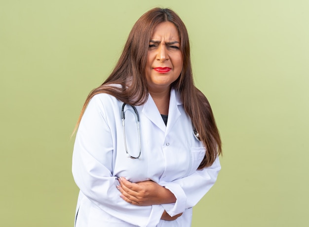 Middle age woman doctor in white coat with stethoscope looking unwell touching her bely feeling pain standing over green wall