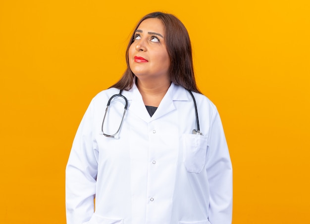 Middle age woman doctor in white coat with stethoscope looking up puzzled