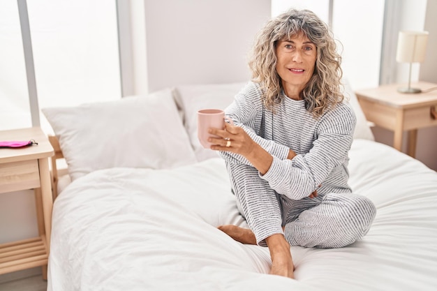 Middle age woman drinking cup of coffee sitting on bed at bedroom