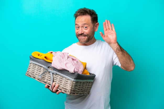 Middle age woman holding a clothes basket isolated on blue background saluting with hand with happy expression