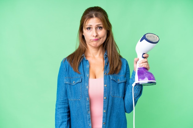 Middle age woman holding an iron over isolated background with sad expression