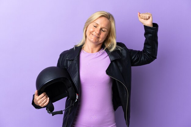 Middle age woman holding a motorcycle helmet isolated on purple wall doing strong gesture