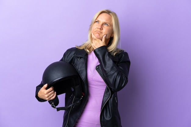 Middle age woman holding a motorcycle helmet isolated on purple wall having doubts