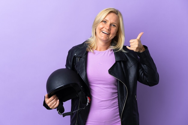 Middle age woman holding a motorcycle helmet isolated on purple wall making phone gesture. Call me back sign