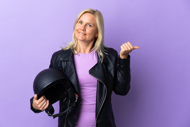 Middle age woman holding a motorcycle helmet isolated on purple wall pointing to the side to present a product