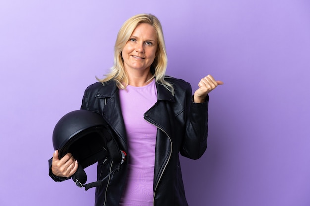 Middle age woman holding a motorcycle helmet isolated on purple wall pointing to the side to present a product