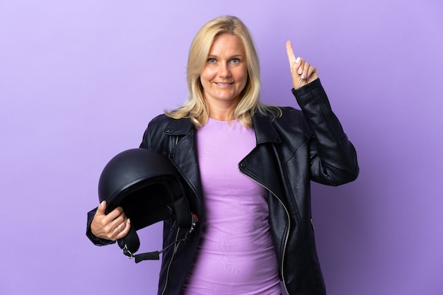 Middle age woman holding a motorcycle helmet isolated on purple wall pointing up a great idea