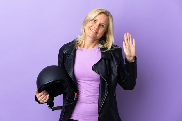 Middle age woman holding a motorcycle helmet isolated on purple wall saluting with hand with happy expression