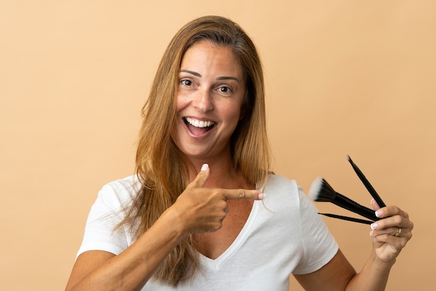 Middle age woman isolated on beige wall holding makeup brush and pointing it