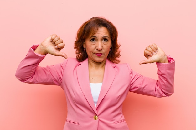 Middle age woman looking sad, disappointed or angry, showing thumbs down in disagreement, feeling frustrated against pink wall