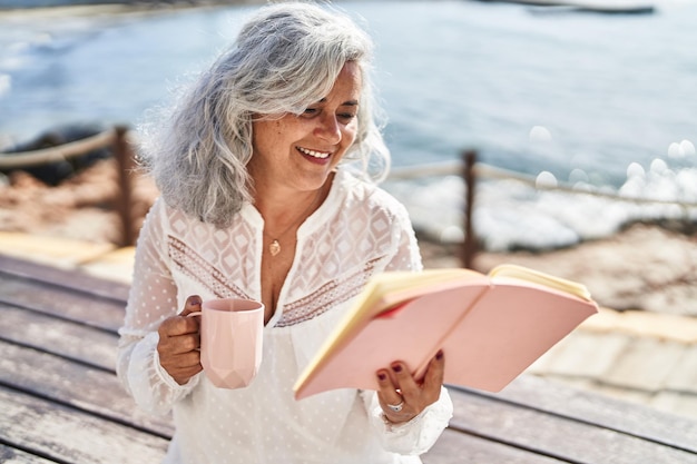 Middle age woman reading book and drinking coffee sitting on bench at seaside