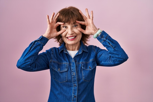 Middle age woman standing over pink background trying to open eyes with fingers, sleepy and tired for morning fatigue