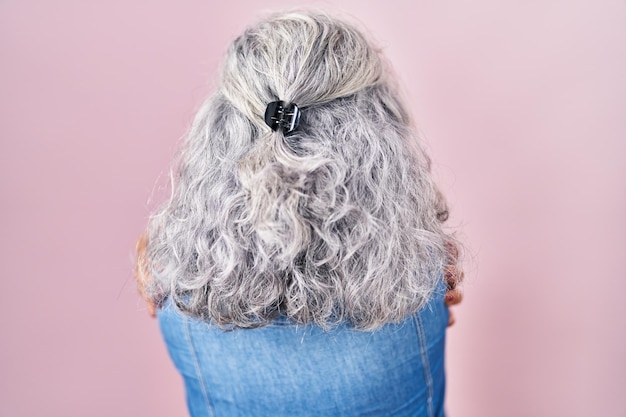 Middle age woman with grey hair standing over pink background hugging oneself happy and positive from backwards self love and self care