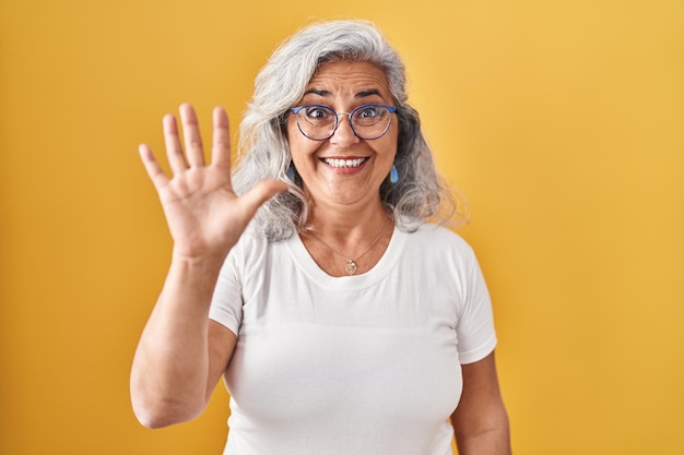 Middle age woman with grey hair standing over yellow background showing and pointing up with fingers number five while smiling confident and happy.