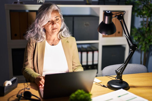 Middle age woman with grey hair working using computer laptop late at night relaxed with serious expression on face. simple and natural looking at the camera.