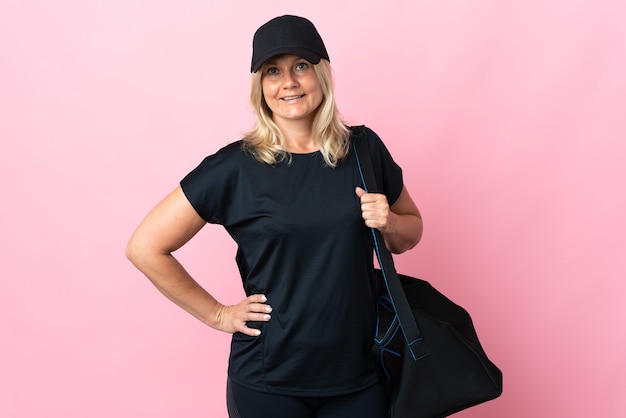 Middle age woman with sport bag isolated on pink wall posing with arms at hip and smiling